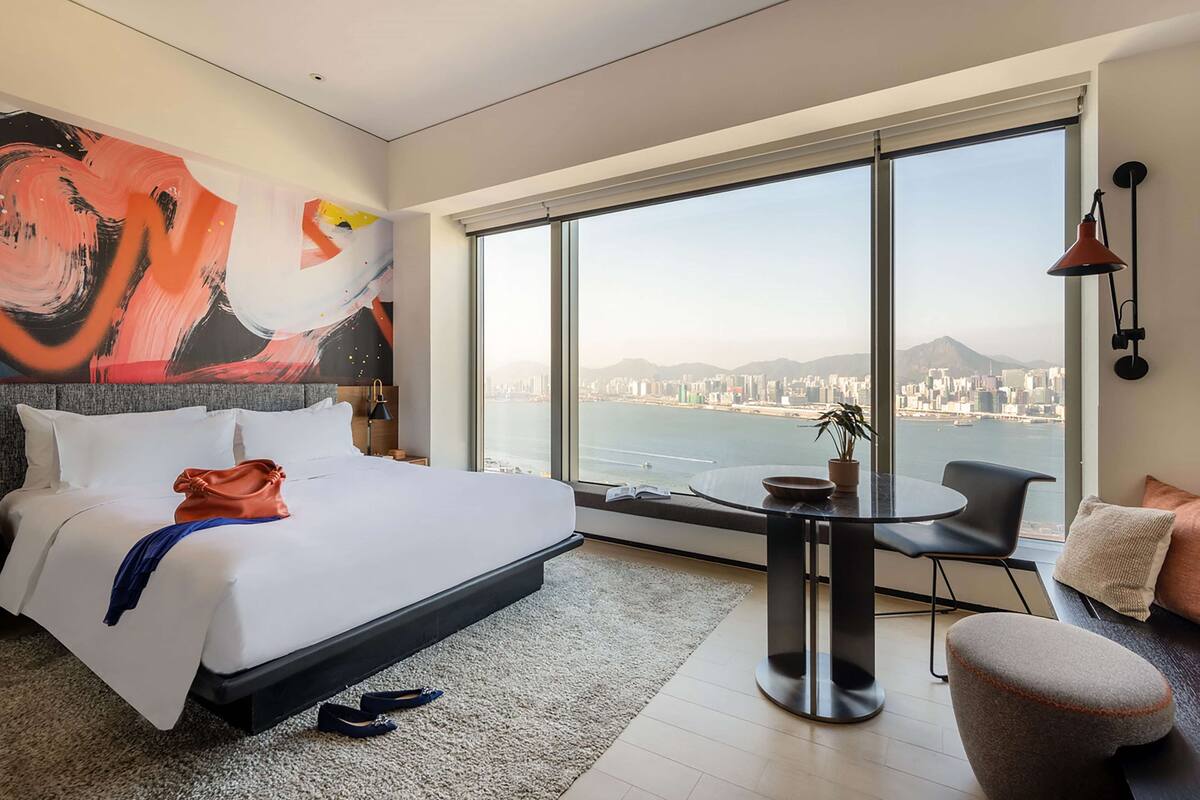 A modern hotel room with a large bed, a round table, and a view of a cityscape through large windows.