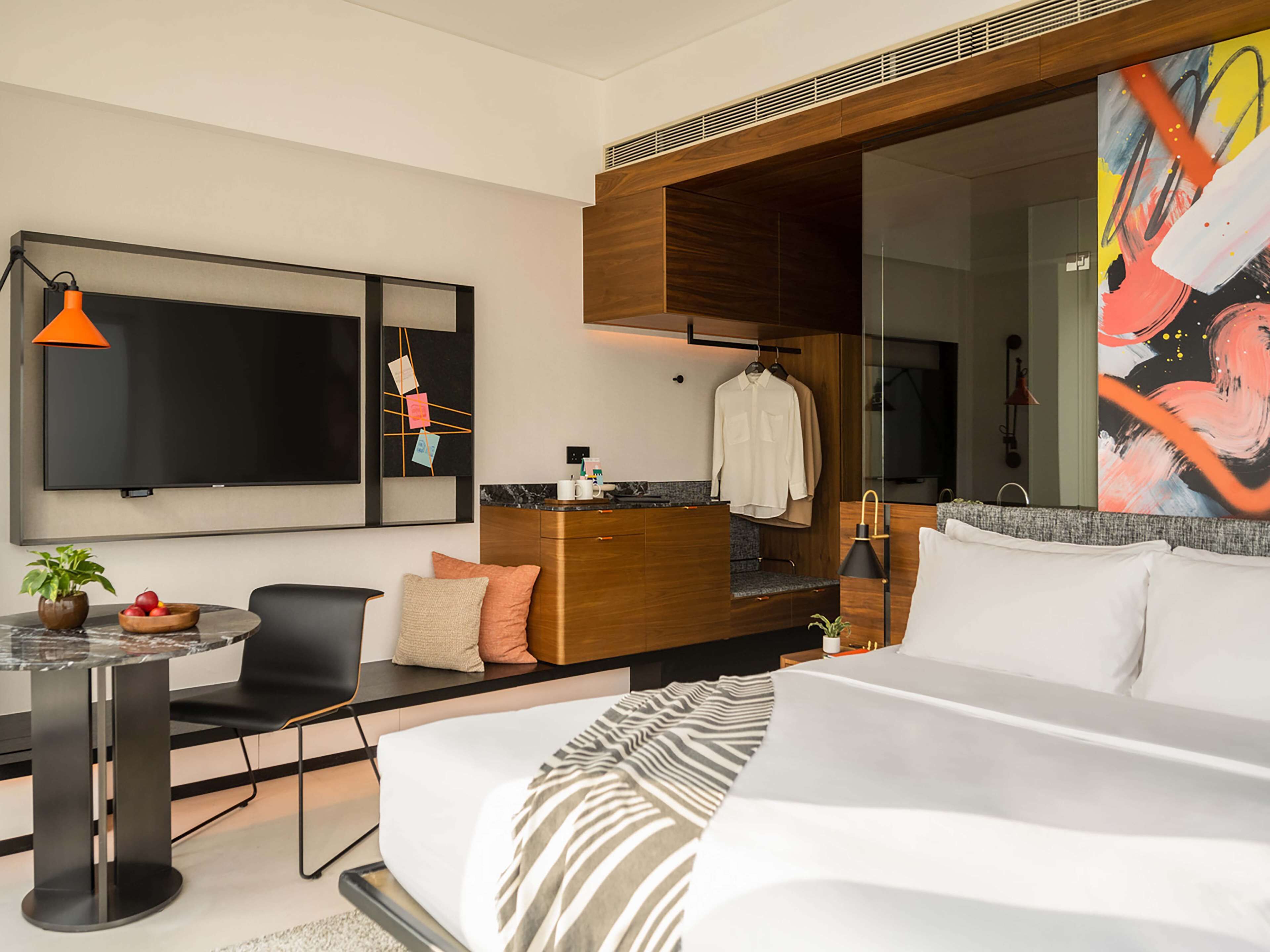 A modern hotel room with a large bed, a desk with a chair, a TV, a painting, and a bathroom visible through a glass partition.