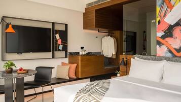 A modern hotel room with a large bed, a desk with a chair, a TV, a painting, and a bathroom visible through a glass partition.