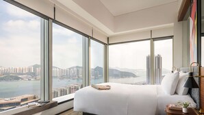 Ein modernes Hotelzimmer mit einem großen Bett, einem Nachttisch, und Blick auf Stadt und Wasser durch raumhohe Fenster.