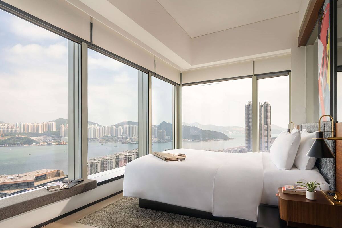 A modern hotel room with a large bed, a bedside table, a nightstand, and a view of a cityscape and water through floor-to-ceiling windows.