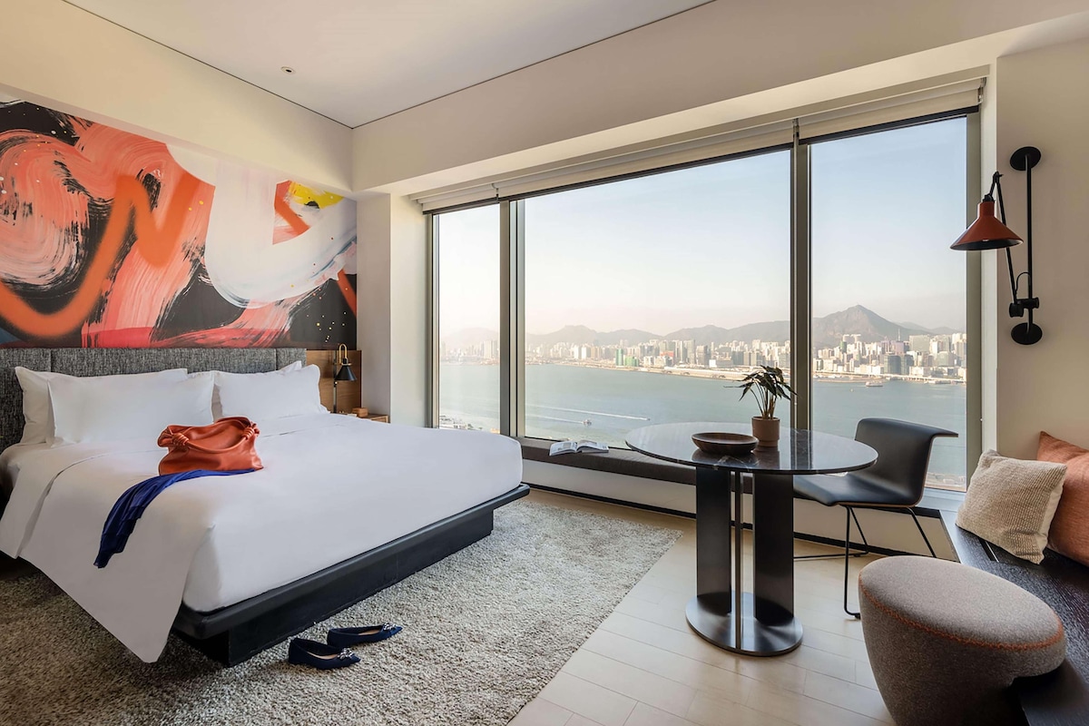 A modern hotel room with a large bed, a round table, a chair, and a view of a cityscape through large windows.