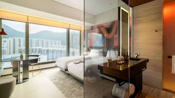 Une chambre d’hôtel moderne dotée d’un grand lit, d’une paroi en verre, d’une salle de bains avec miroir et offrant une vue sur le paysage urbain.