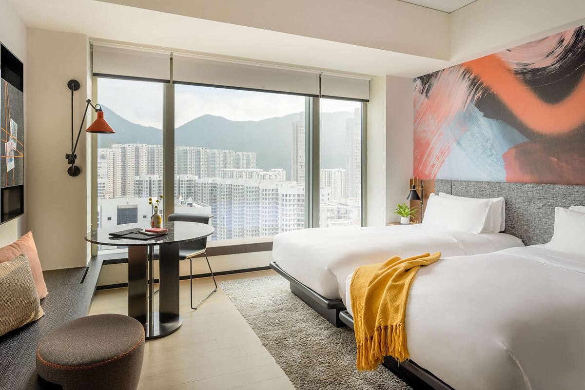 A hotel room with two beds, a desk, a chair, a large window with a city view, and a painting on the wall.
