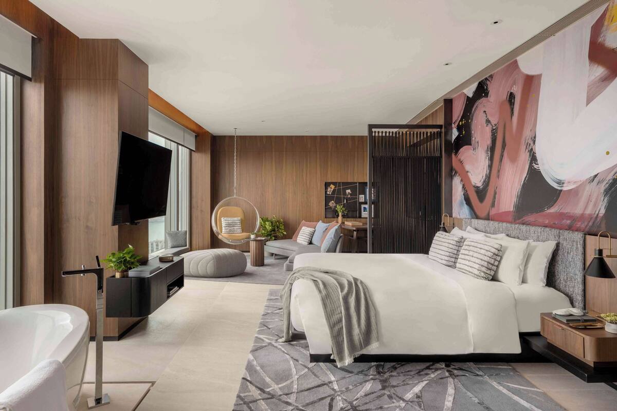 A modern hotel room with a large bed, a sitting area with a sofa and armchairs, a television, and a view of the city.