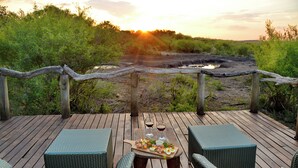 An outdoor deck with wicker furniture and a table set for a meal, overlooking a natural landscape with a sunset.