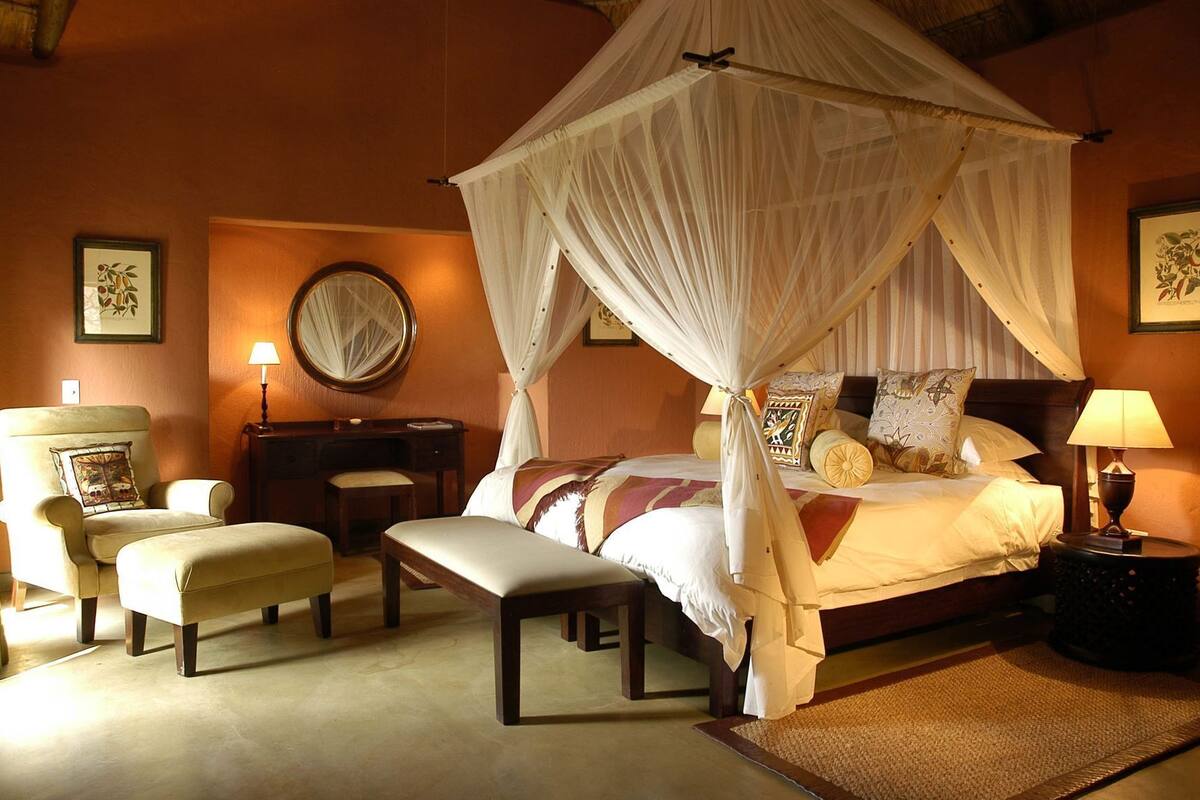 Een slaapkamer met hemelbed, een zithoek met een stoel en een bankje, een nachtkastje met lamp en een spiegel aan de muur.