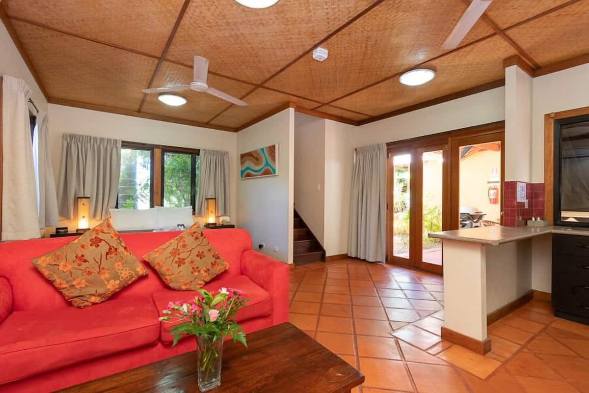 A living room with a red sofa, a wooden coffee table, and a vase of flowers. There is a staircase leading to an outdoor area.