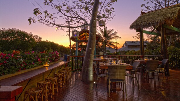 An outdoor dining area with wooden tables and chairs, surrounded by lush greenery and a thatched-roof structure. - Bali Hai Resort & Spa (Cable Beach)