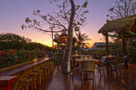 An outdoor dining area with wooden tables and chairs, surrounded by lush greenery and a thatched-roof structure.
