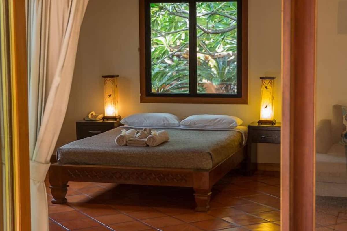 A bedroom with a bed, two bedside tables, a window with a view of greenery, and a decorative ceiling panel.