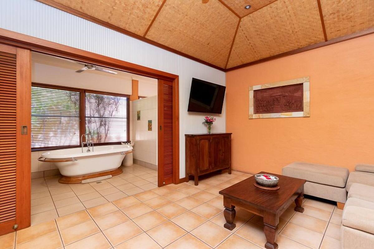 A bathroom with a bathtub, a toilet, and a sink. There is a wooden cabinet, a television mounted on the wall, and a framed picture. The room has a tiled floor and a ceiling with wooden beams.
