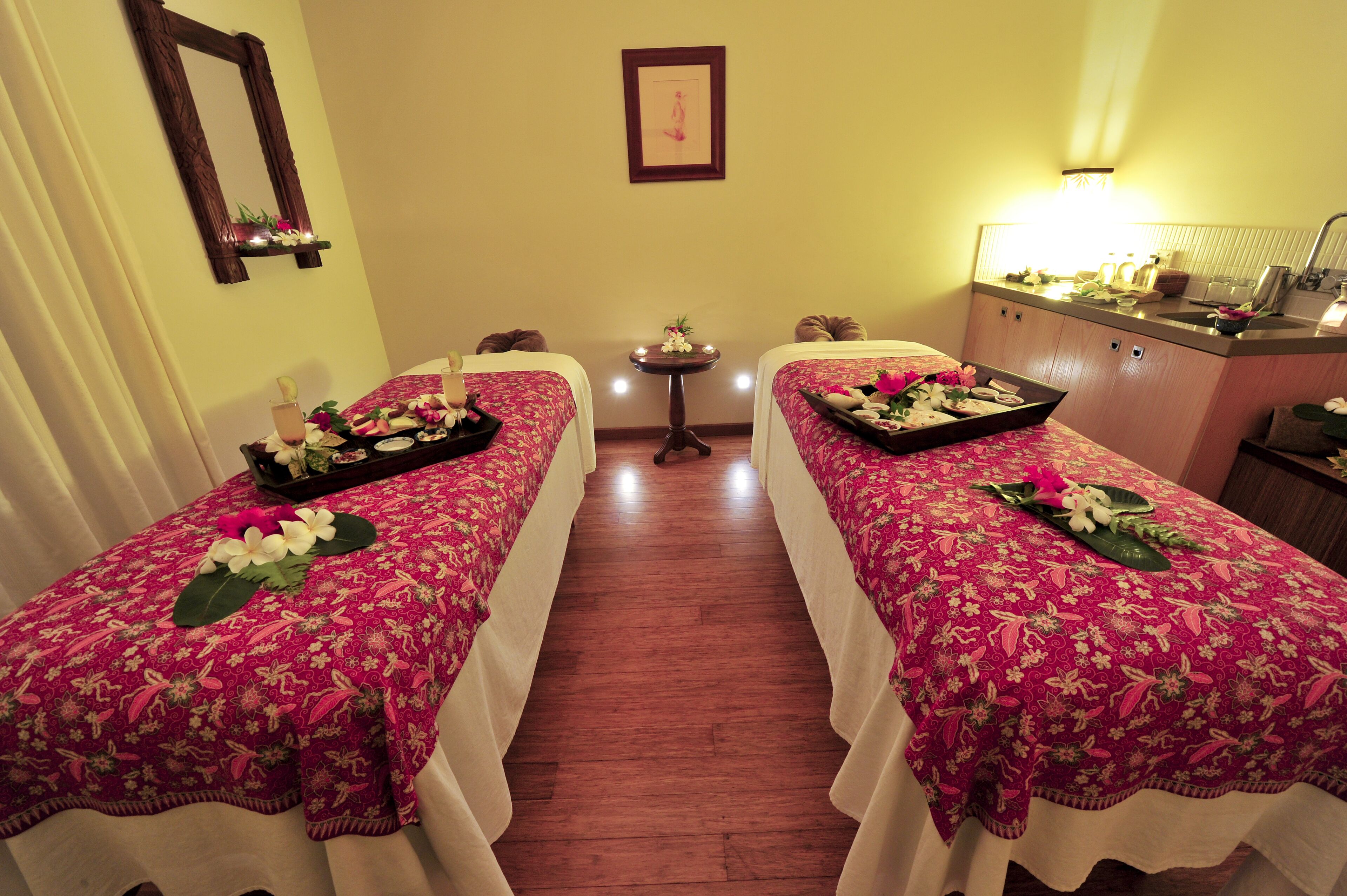 A room with two beds, a small table with flowers, a mirror, and a sink.