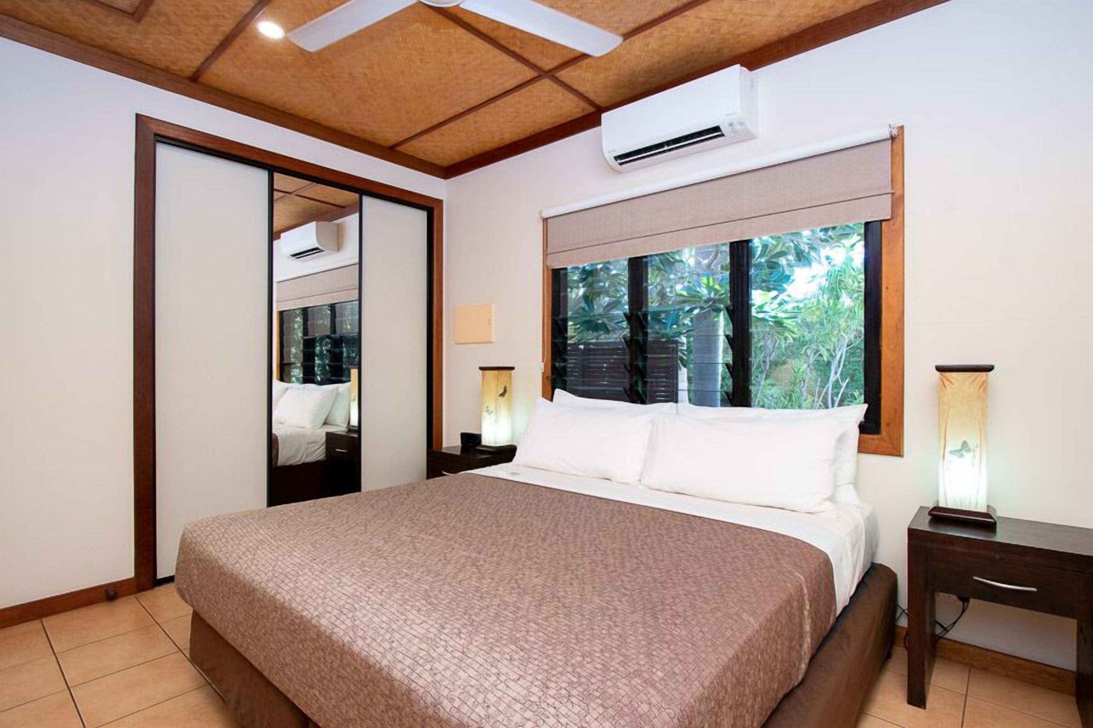 Un dormitorio con una cama grande, una mesita de noche, una lámpara y una ventana que da a vistas de vegetación.