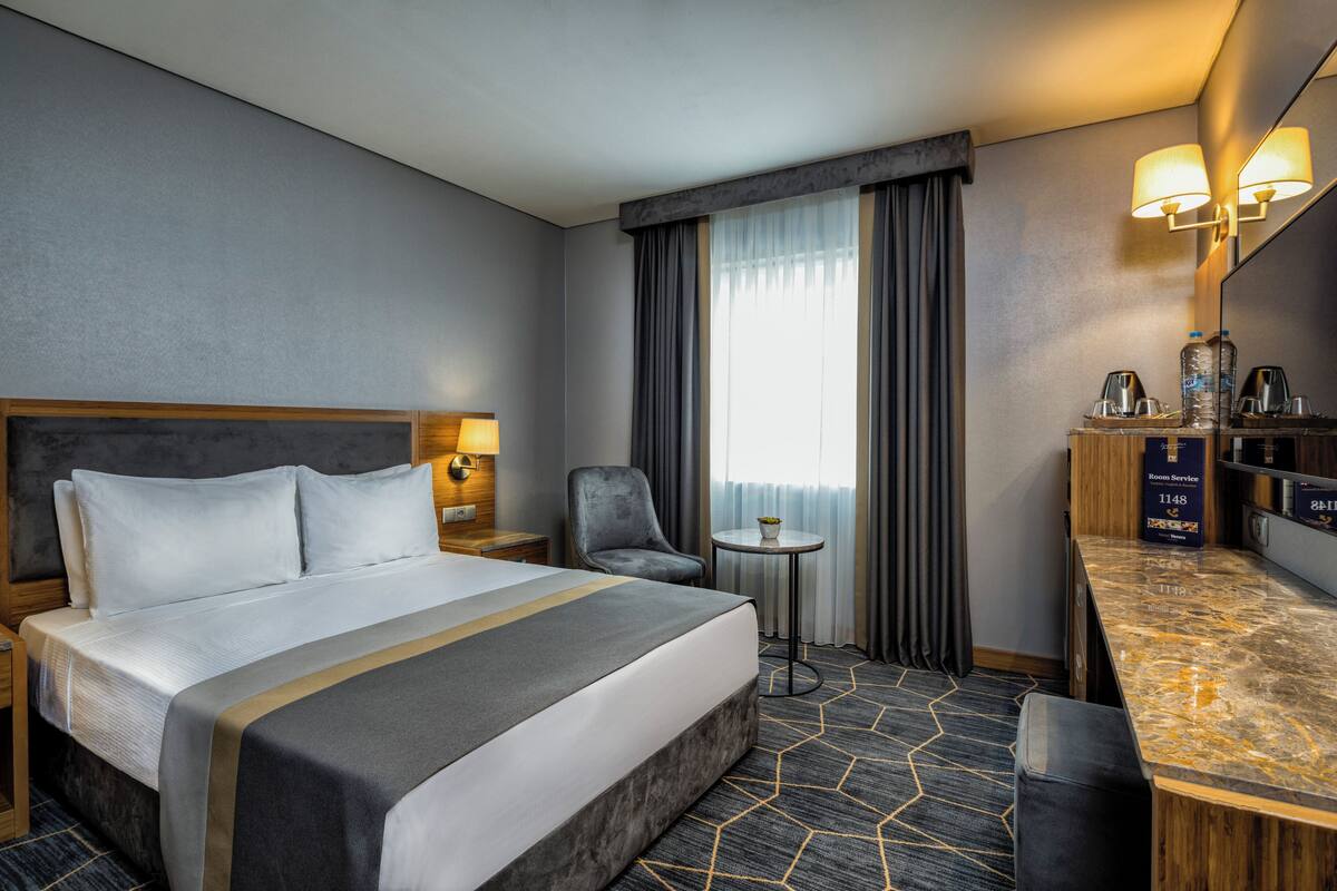 A hotel room with a large bed, a desk with a mirror, a chair, a small table, and a window with curtains.