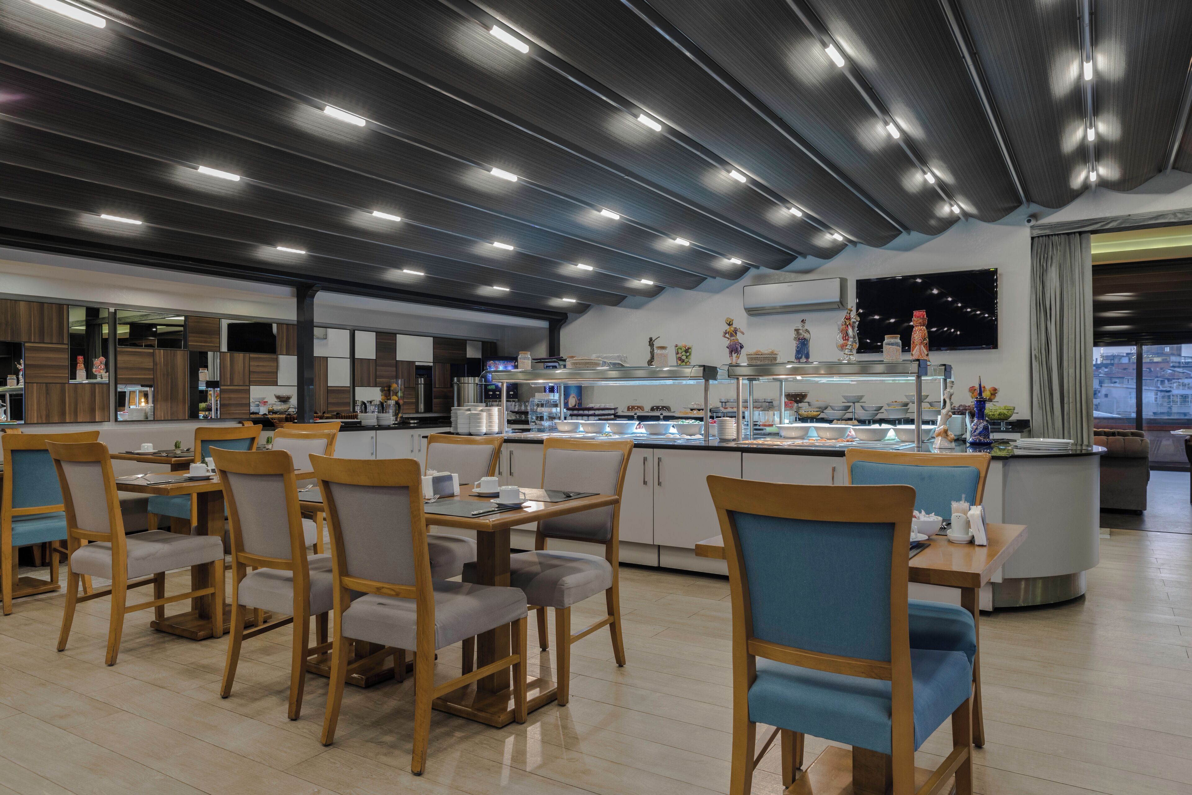 Un restaurante moderno con mesas y sillas de madera, una barra central con mostradores de comida y un amplio ventanal con vistas a la ciudad.