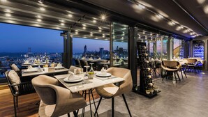 Un restaurante moderno con vistas a la ciudad, con mesas y sillas, una estantería para vinos y un amplio ventanal con vista al paisaje urbano.