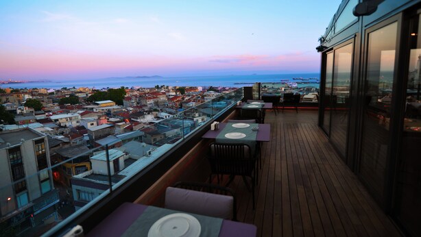 Terraza en la azotea con mesas y sillas, con vistas a un paisaje urbano y al mar al atardecer.