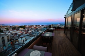 Terraza en la azotea con mesas y sillas, con vistas a un paisaje urbano y al mar al atardecer.