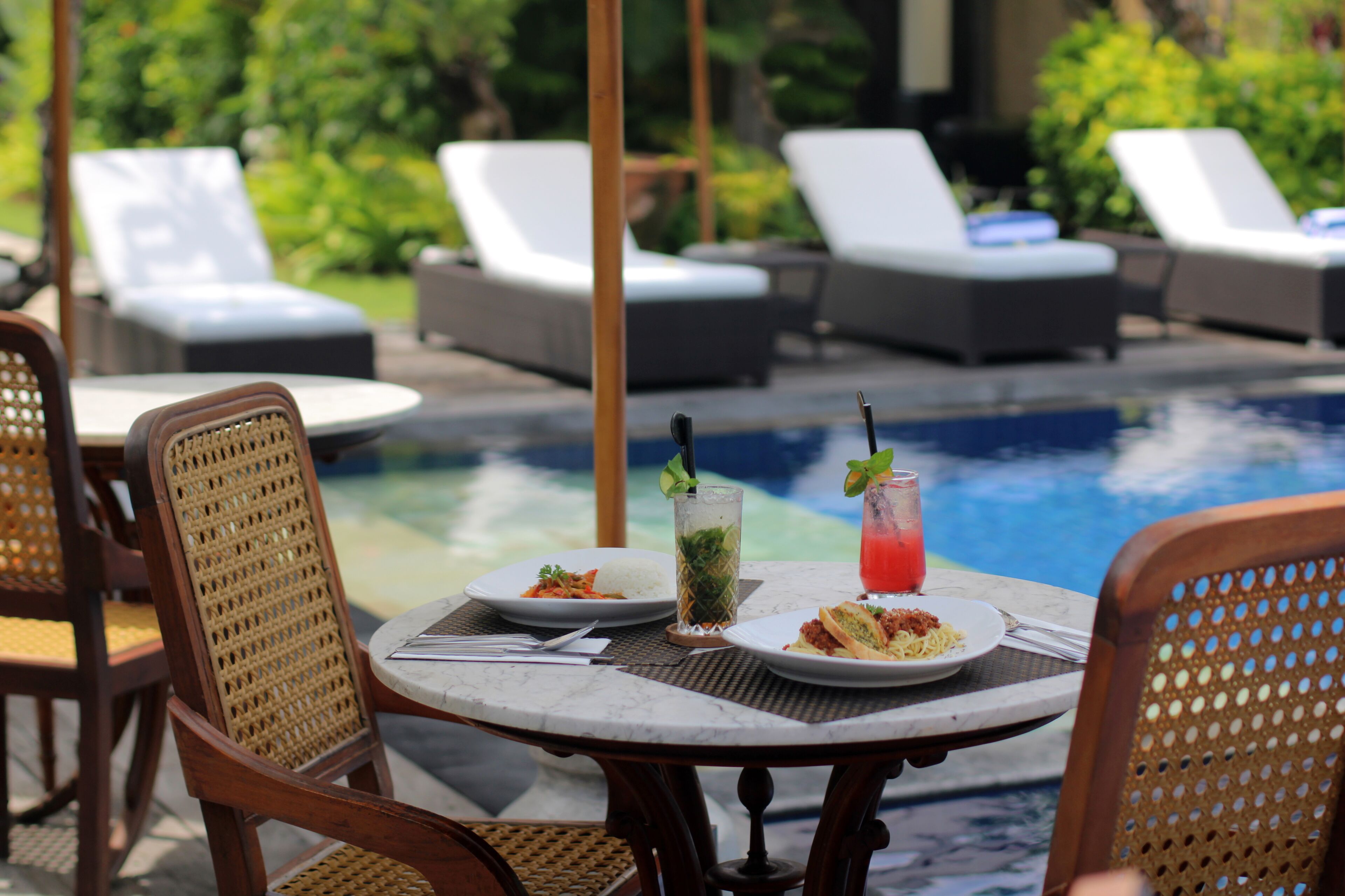A poolside dining area with lounge chairs, a table with plates of food, and a drink.