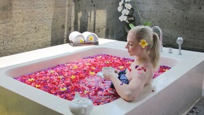 Uma mulher em banheira cheia de pétalas de flores, uma estátua e um porta-toalhas.