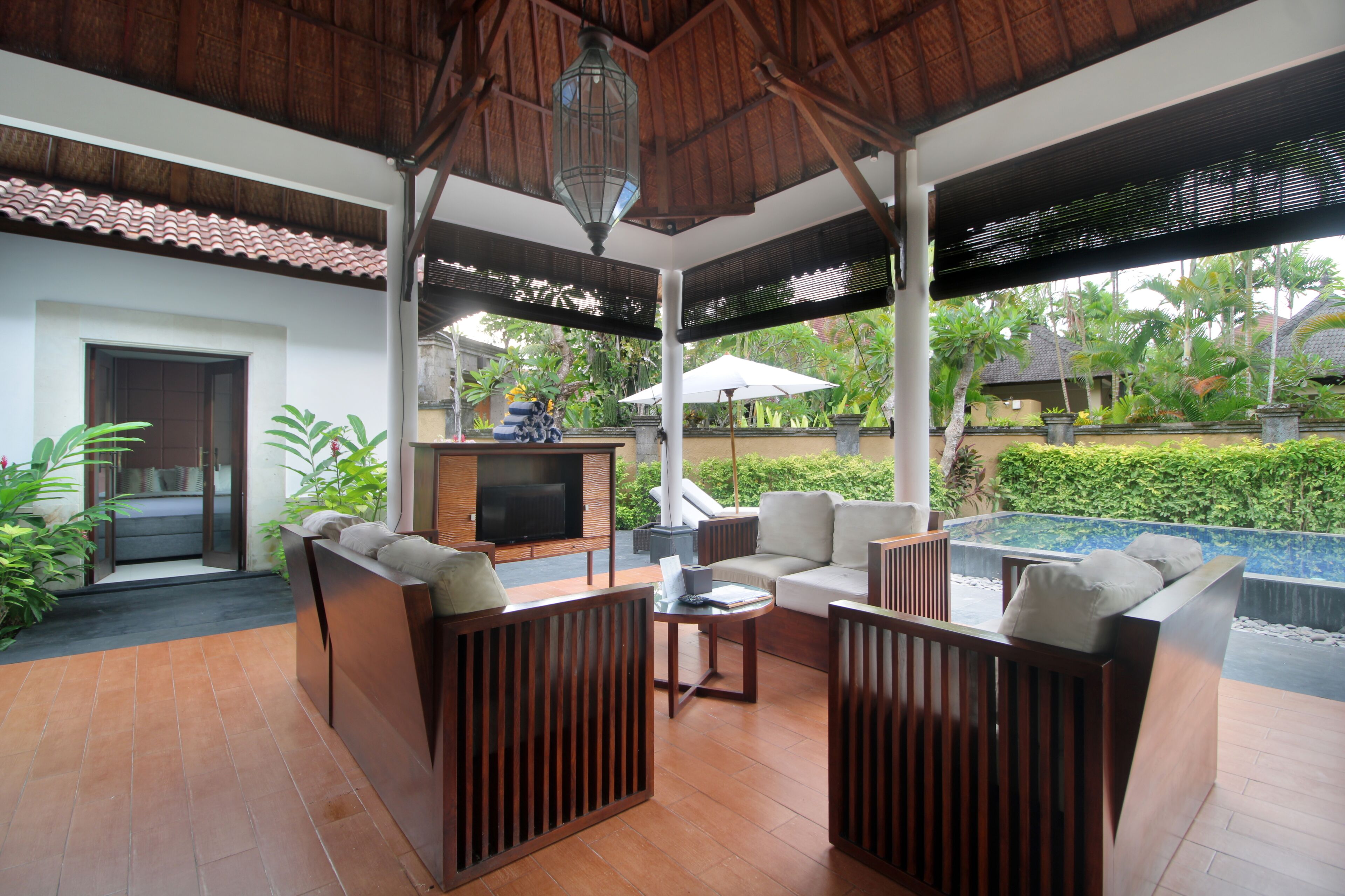 A covered outdoor seating area with wooden furniture, a television, and a pool in the background.