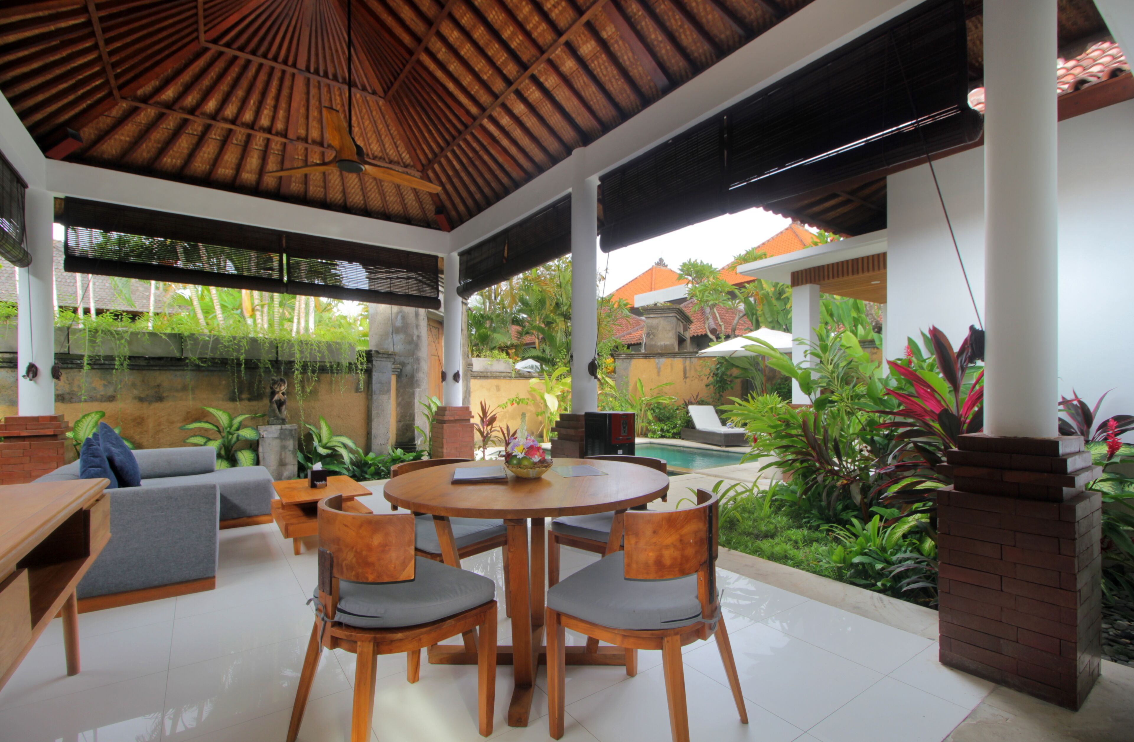 A covered outdoor seating area with a round table and chairs, surrounded by lush greenery and a view of a swimming pool.