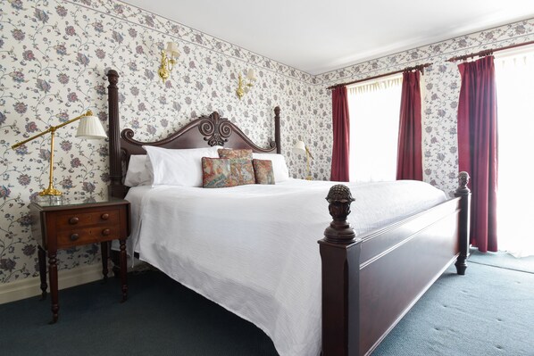 A bedroom with a four-poster bed, floral wallpaper, a nightstand with a lamp, and a window with red curtains. - The Waring House (Prince Edward)