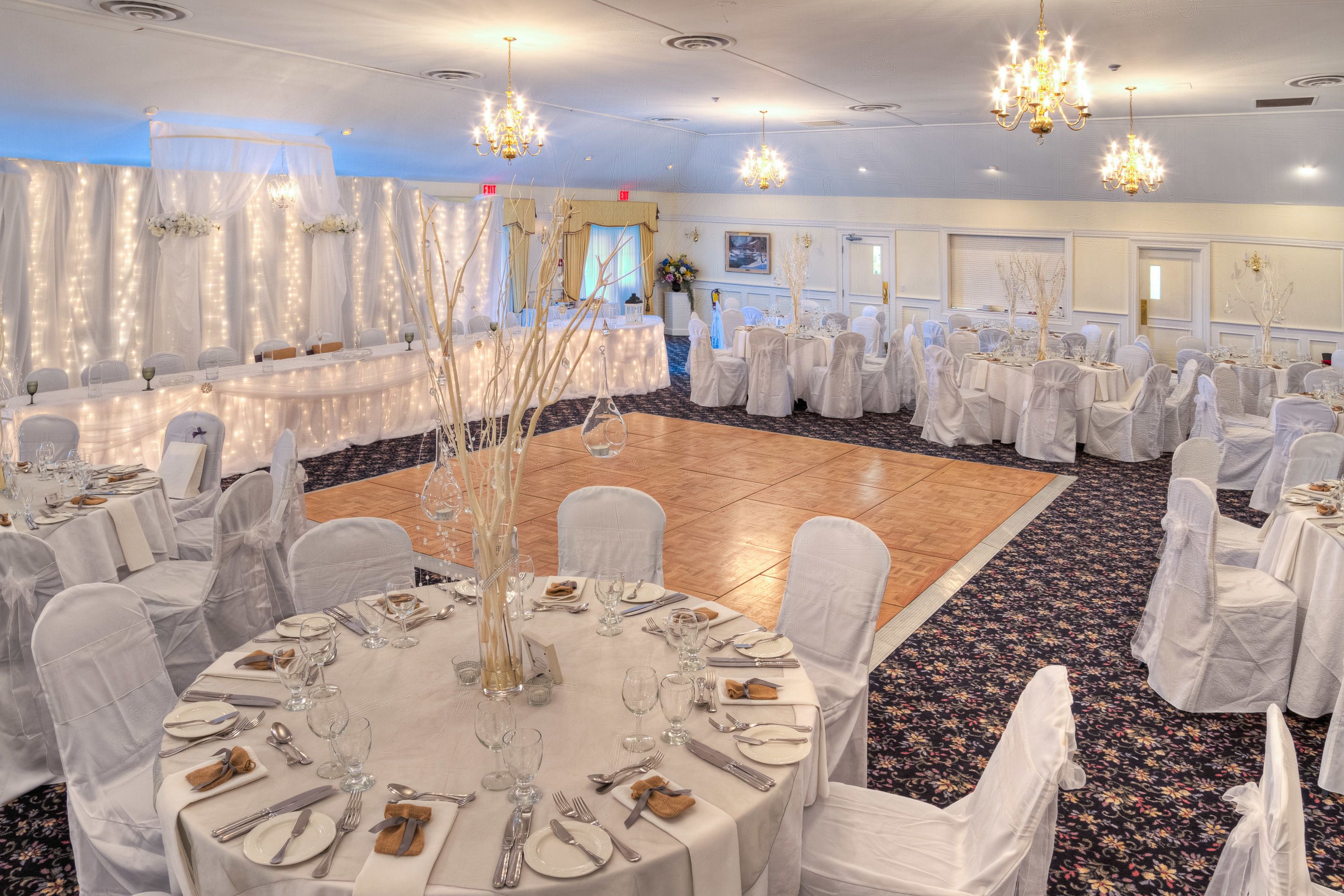 A banquet hall with round tables set for dining, white chairs, chandeliers, and a dance floor.