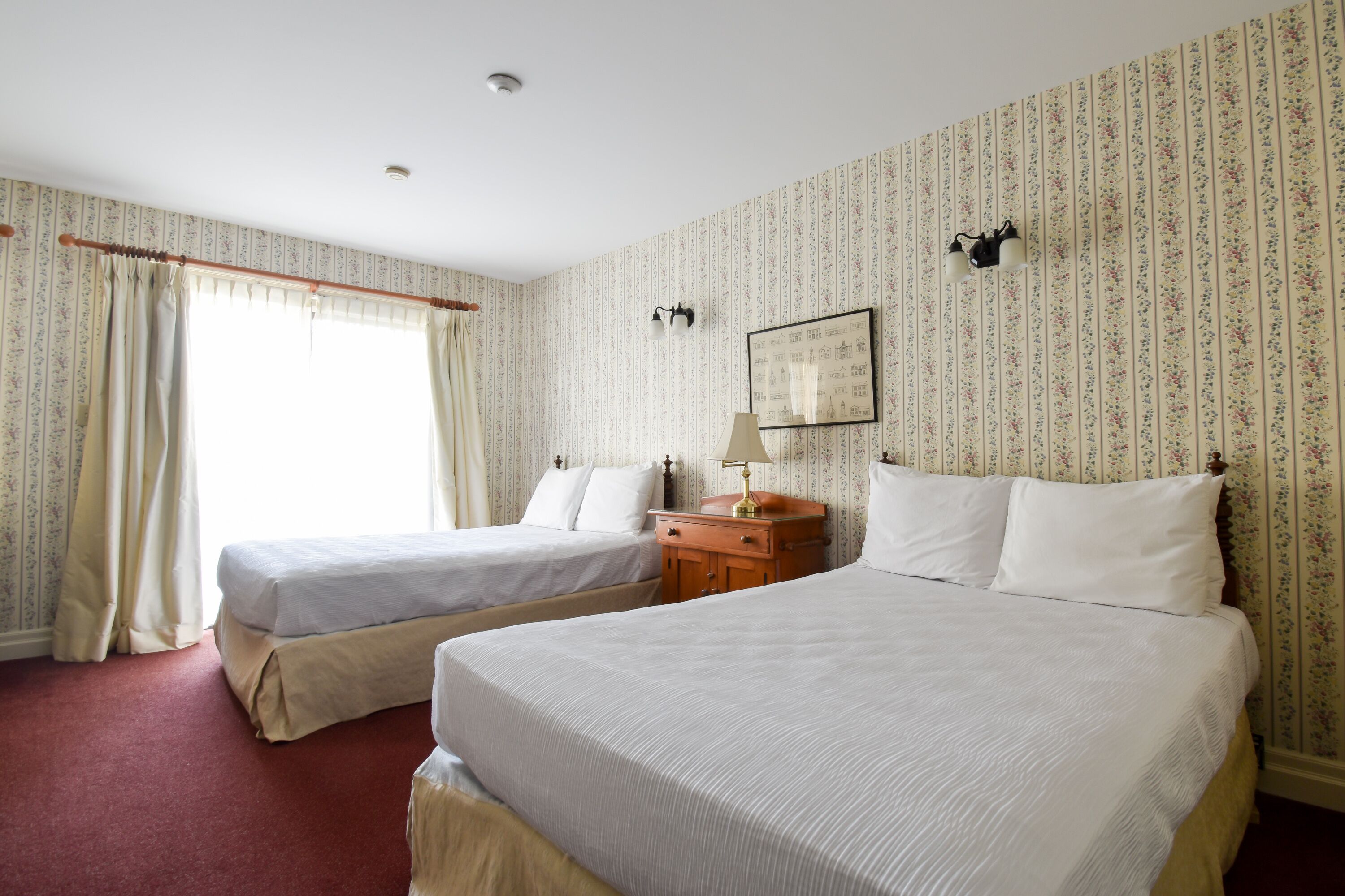 Habitación de hotel con dos camas individuales, una mesita de noche de madera, un espejo y una ventana con cortinas.