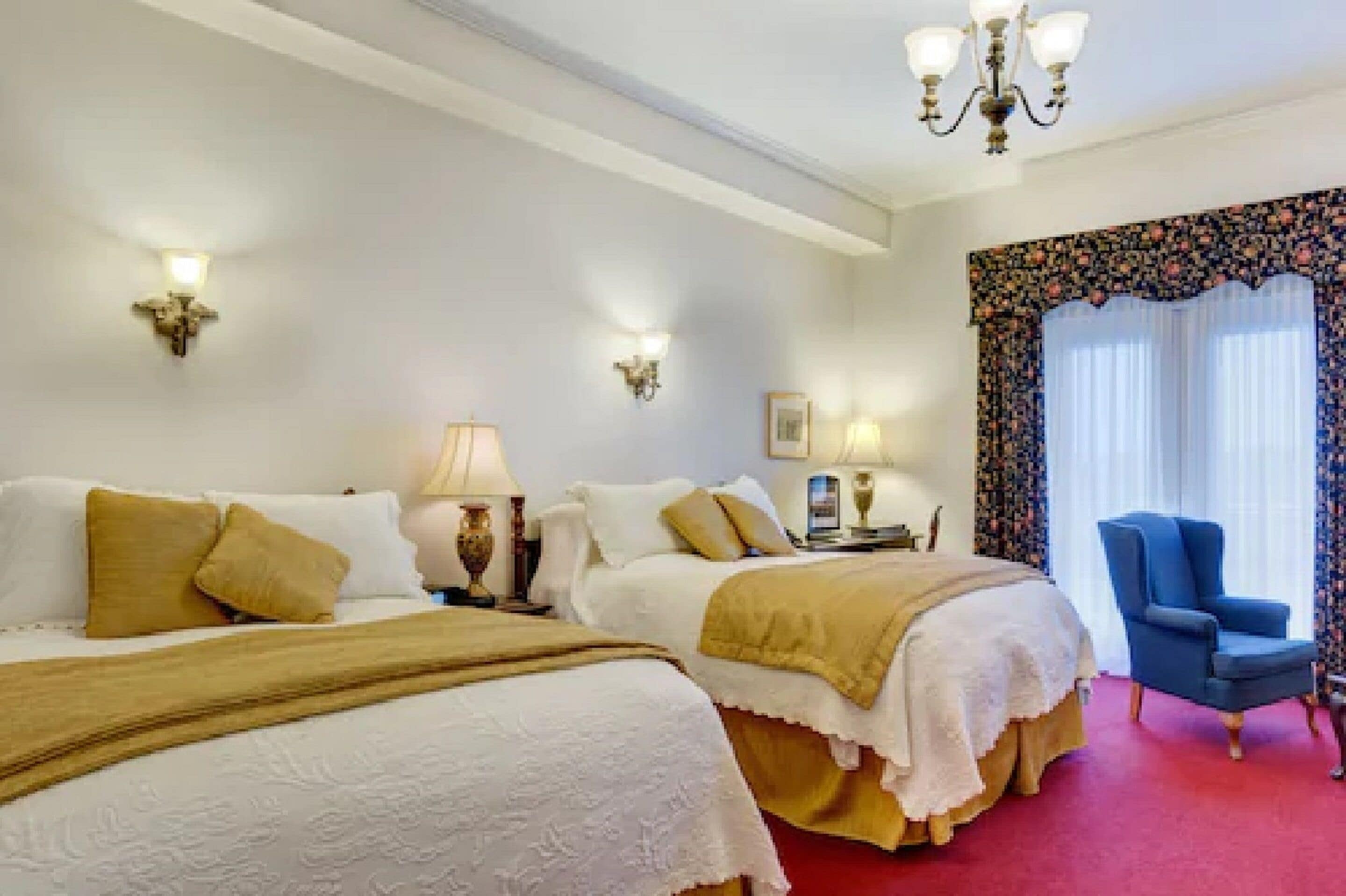 A hotel room with two beds, a blue armchair, and decorative wall sconces.