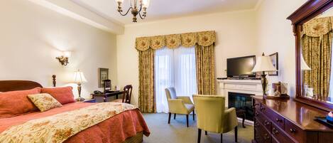 A hotel room with a large bed, a desk, a chair, a dresser, a television, and a fireplace.