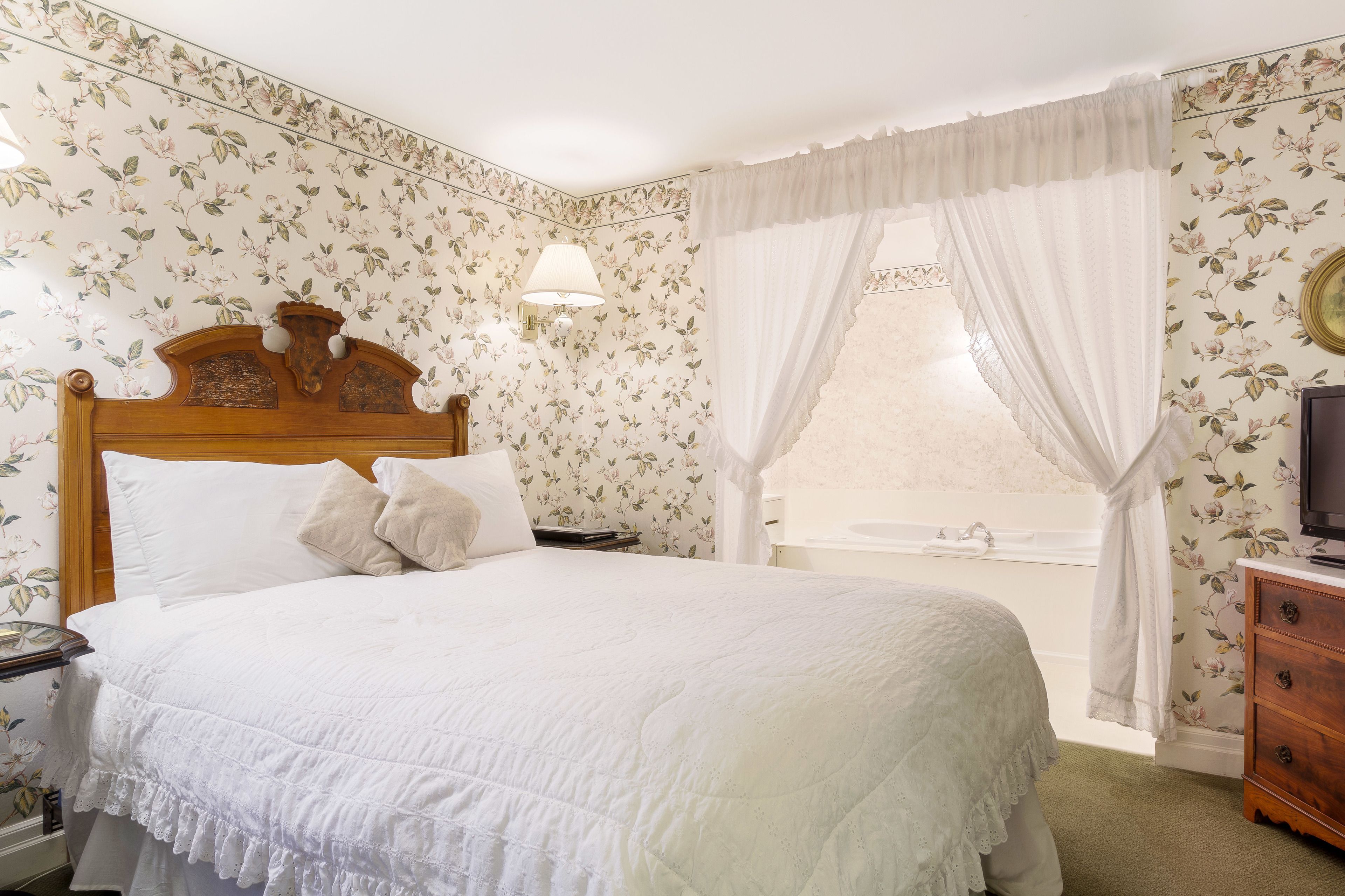 Un dormitorio con cabecera de madera, cubrecama blanco, mesita de noche, lámpara, cómoda y ventana con cortina.