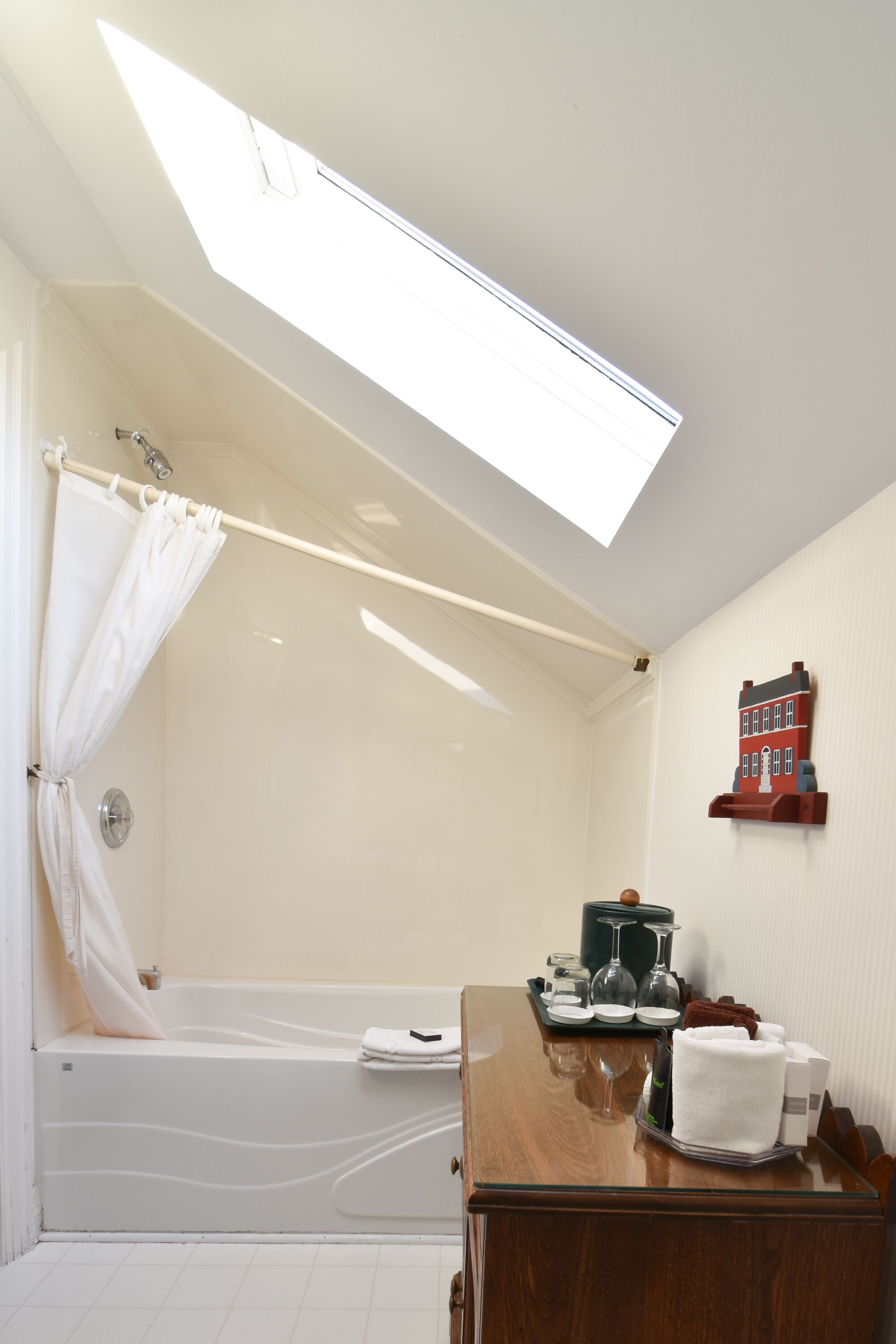 A bathroom with a skylight, a bathtub, a wooden vanity, and a small red house decoration on the wall.
