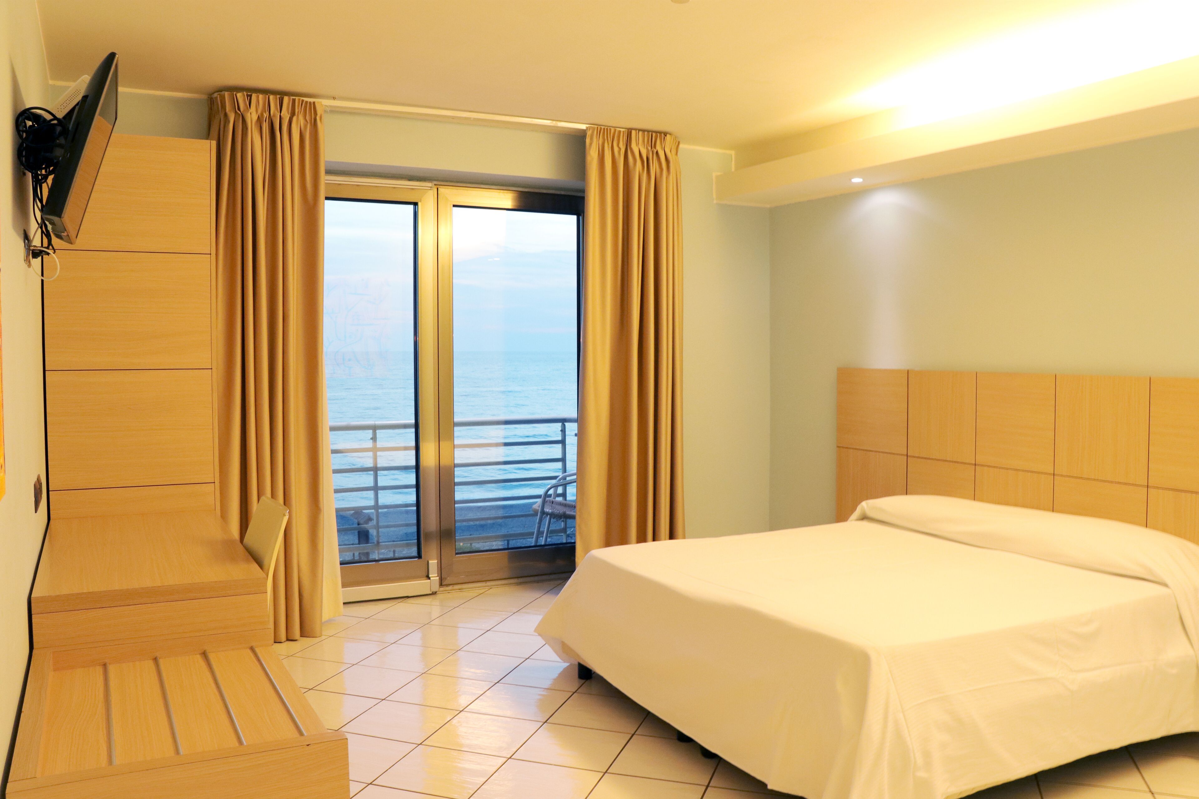 A hotel room with a bed, a TV mounted on the wall, a desk, and a view of the sea through a sliding door.