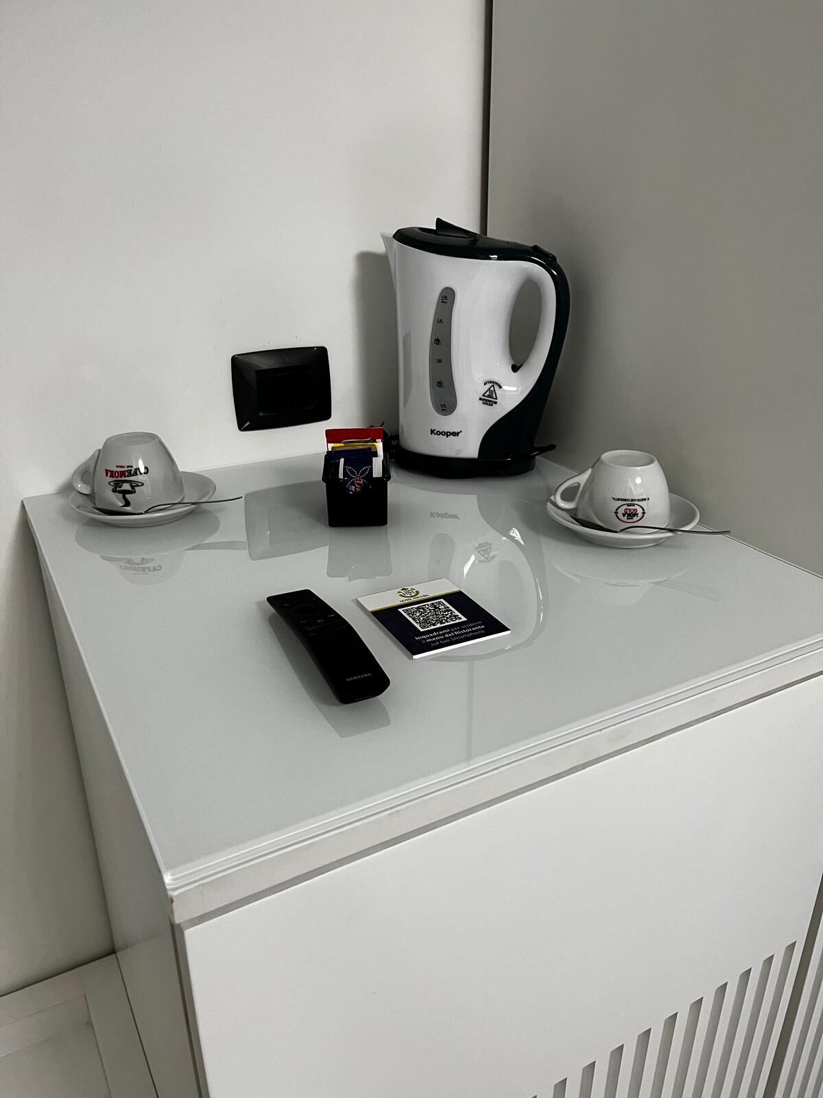 A white minibar with a coffee maker, a remote control, a card with a QR code, and a cup and saucer.