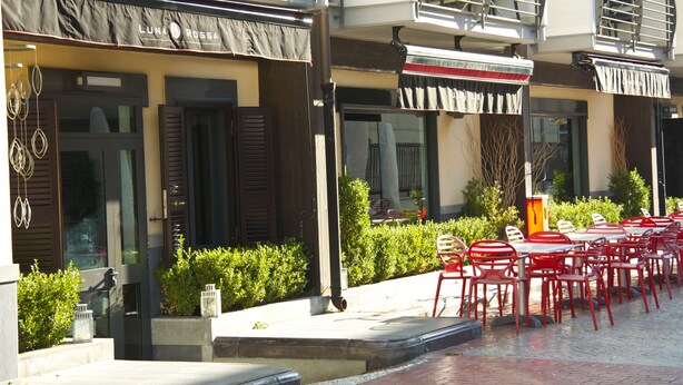 An outdoor seating area with red chairs and tables, a building with a black awning, and greenery. - Rama Palace Hotel (Casalnuovo di Napoli)