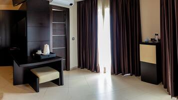 A hotel room with a dark wooden desk, a bench, a small table with a cup and saucer, a TV, and a window with curtains.