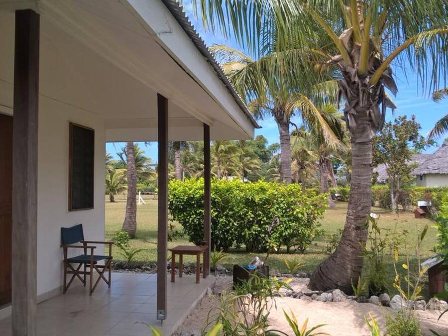 Un porche techado con una silla y una mesa, rodeado de vegetación tropical y un edificio con techo de paja.