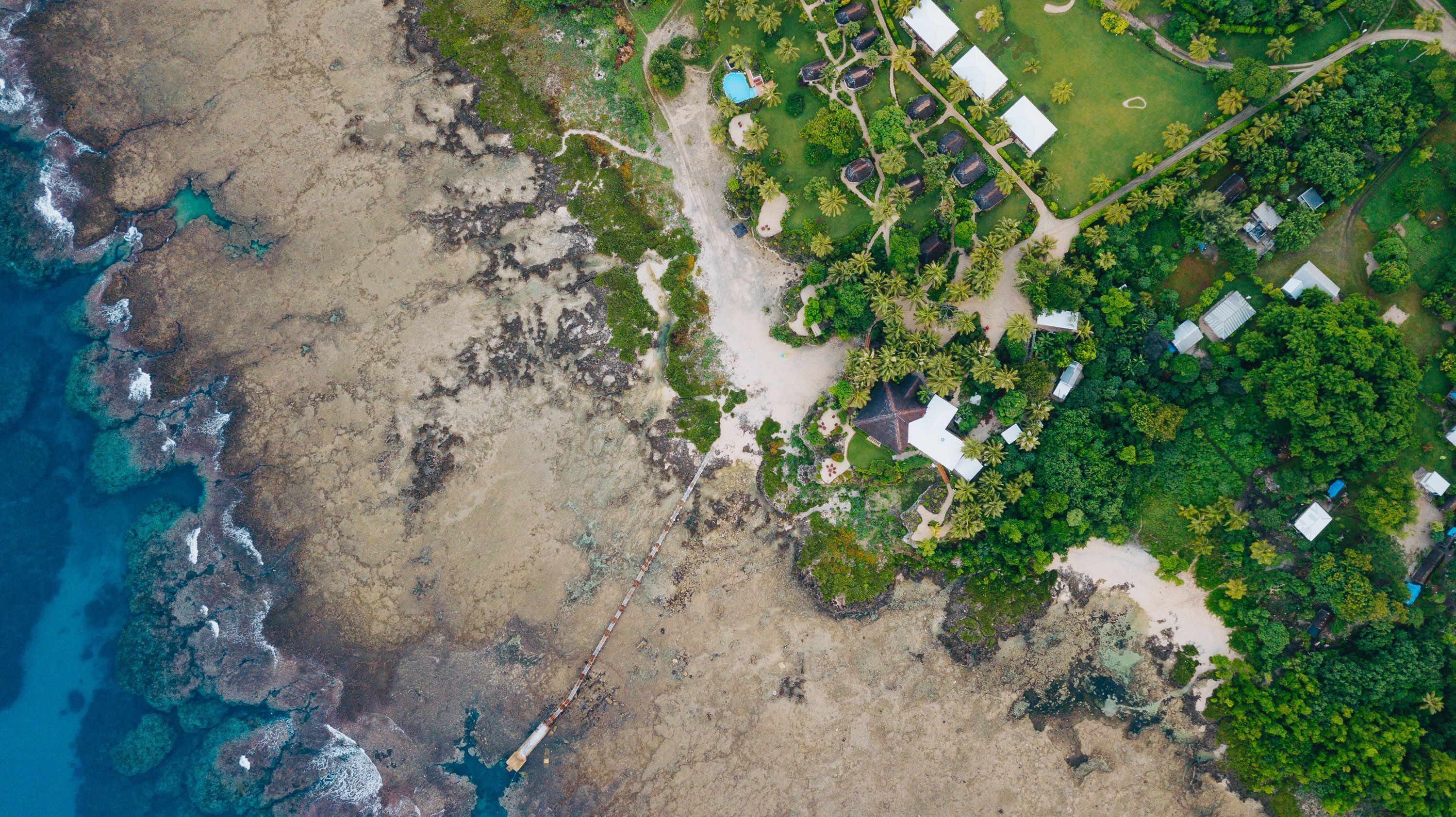 Vista aérea de un complejo turístico costero con varias cabañas, una piscina central y abundante vegetación.