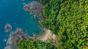 Vista aérea de una zona costera con un bosque verde denso, costa rocosa y agua azul cristalina.