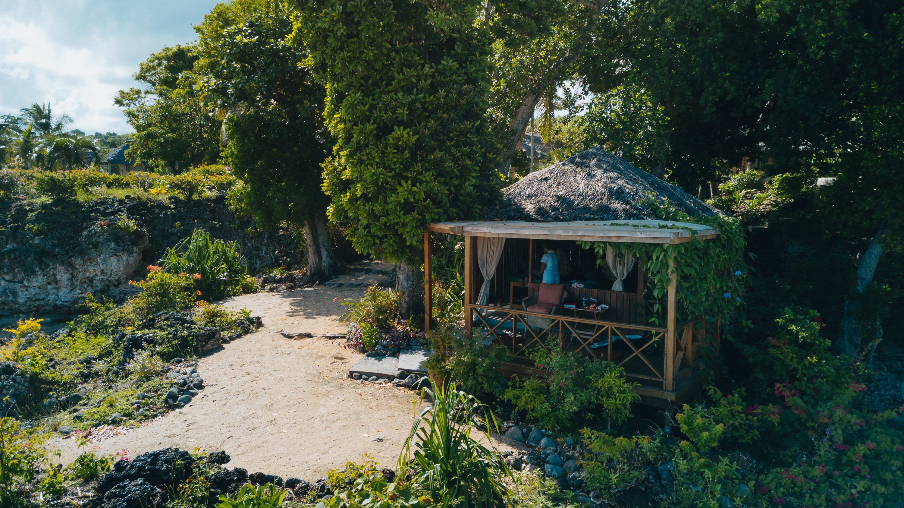 A rustic wooden structure with a thatched roof, surrounded by lush greenery and a dirt path.