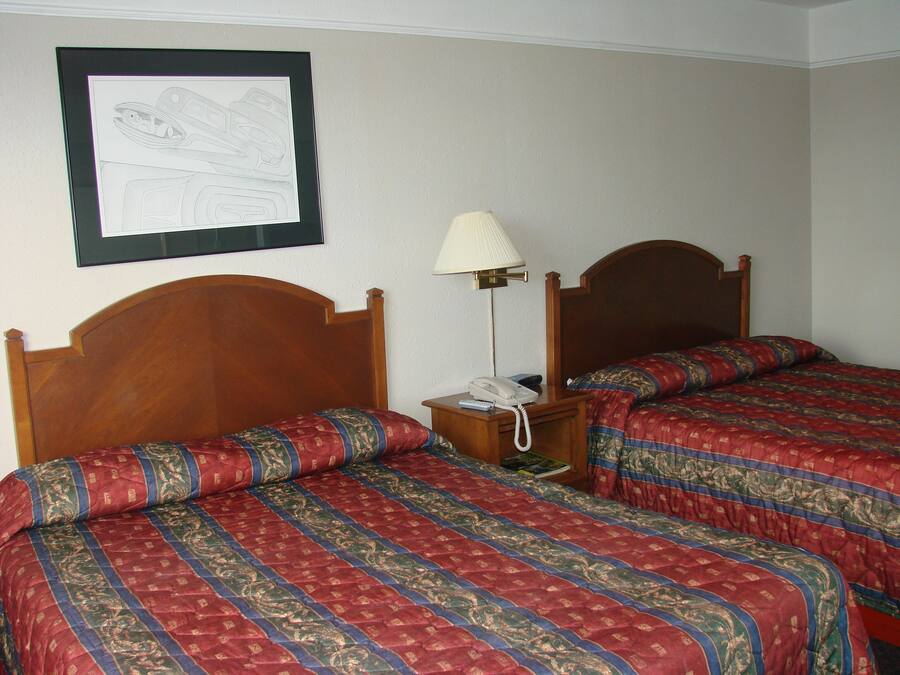 A hotel room with two wooden beds, a bedside table with a lamp, and a framed drawing on the wall.