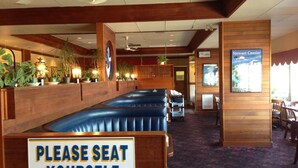 Interior de restaurant amb un cartell que diu 'Please Seat Yourself', seients separats per panells, revestiment de fusta, ventiladors de sostre i un pòster de Stewart Cassiar.
