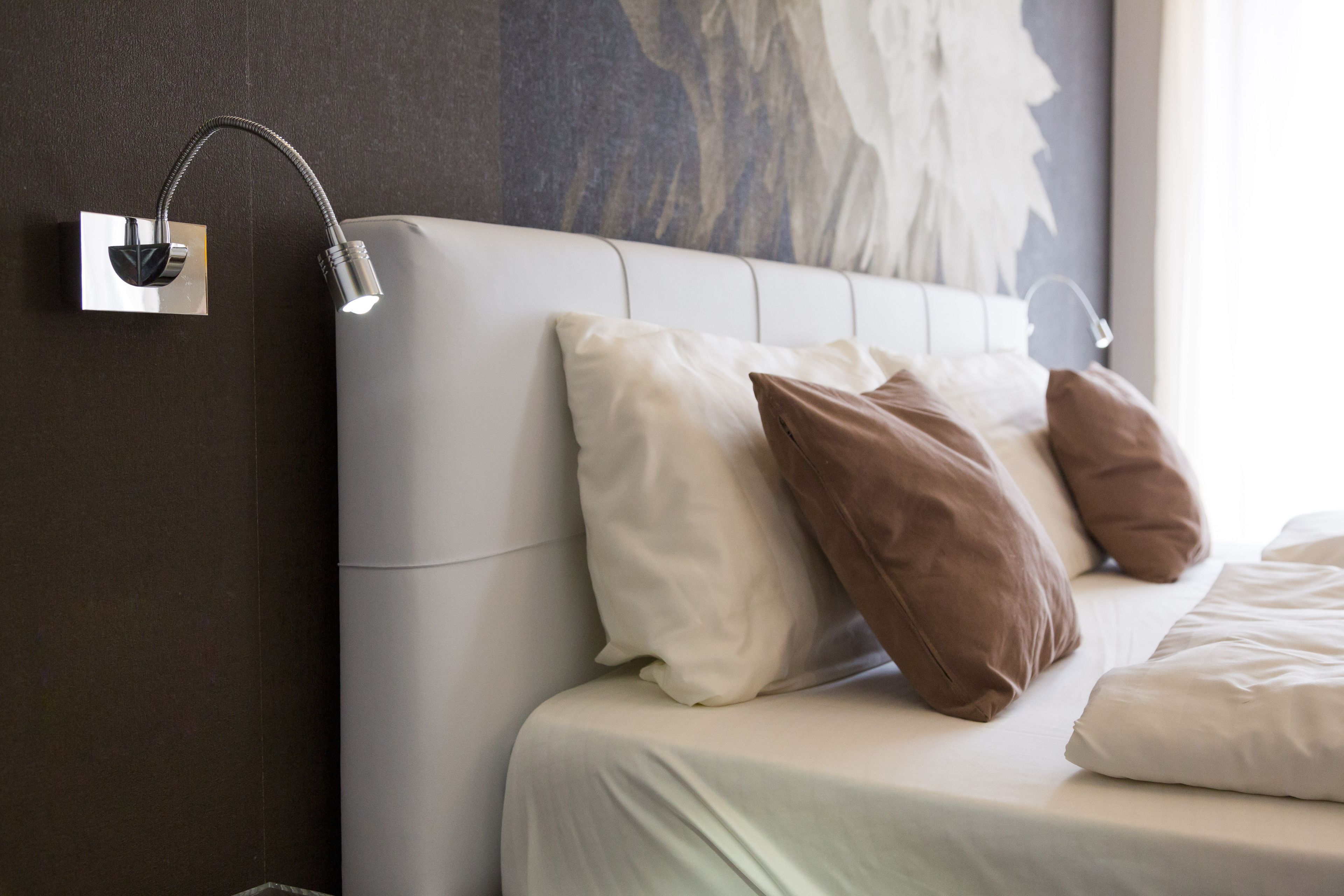 A neatly made bed with white and brown pillows, a modern wall-mounted light fixture, and a large abstract painting on the wall.
