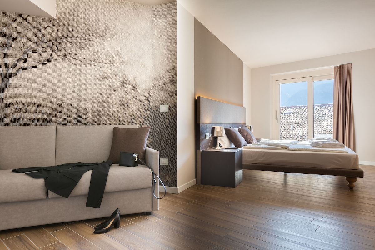 A modern hotel room with a large window, a bed, a sofa, and a wall with a tree mural.