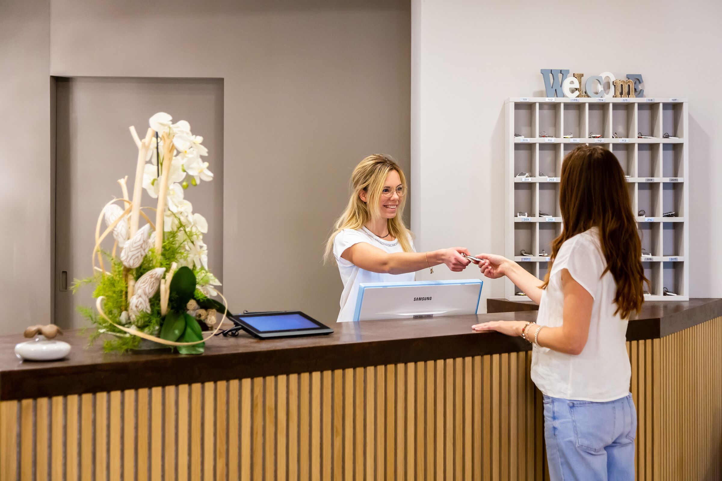 A reception desk with a Samsung monitor, a tablet, and a decorative orchid arrangement.