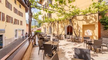 An outdoor café with wicker furniture and a stone wall backdrop, featuring a narrow street and buildings on one side.