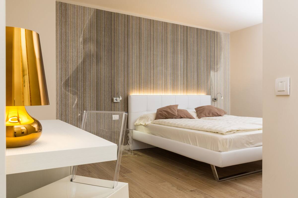 A modern bedroom with a white bed, a clear bedside table, and a striped wallpaper headboard.