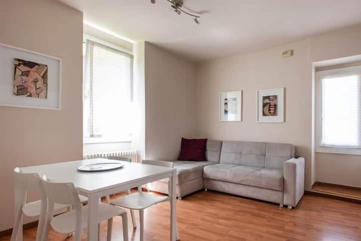 A modern living room with a white sofa, a dining table with chairs, and artwork on the walls.
